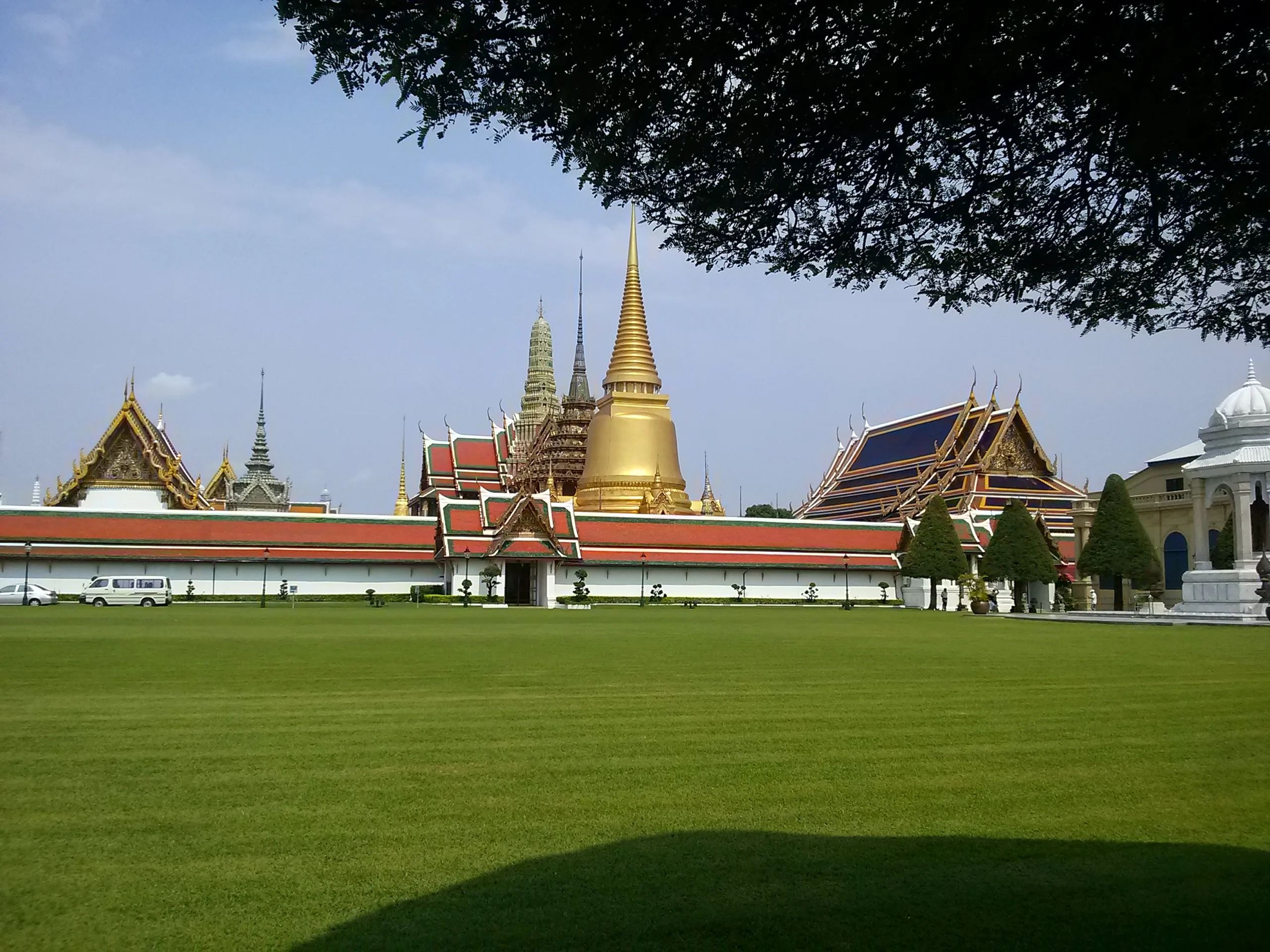 The white outer wall of the Grand Palace seen across the lawn, with golden roofs behind it