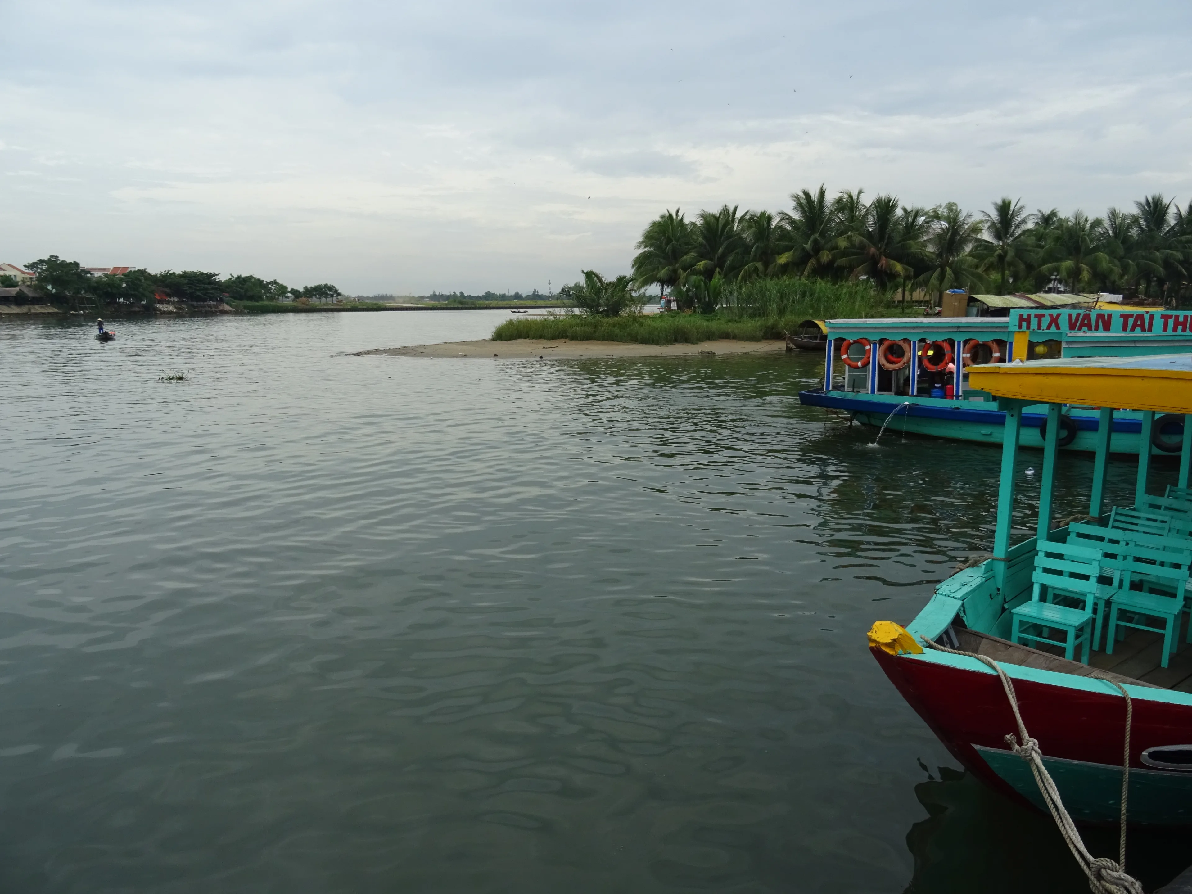 Passenger boats on the Thu Bon at Hoi An, seen from the water’s edge with palms on the far bank