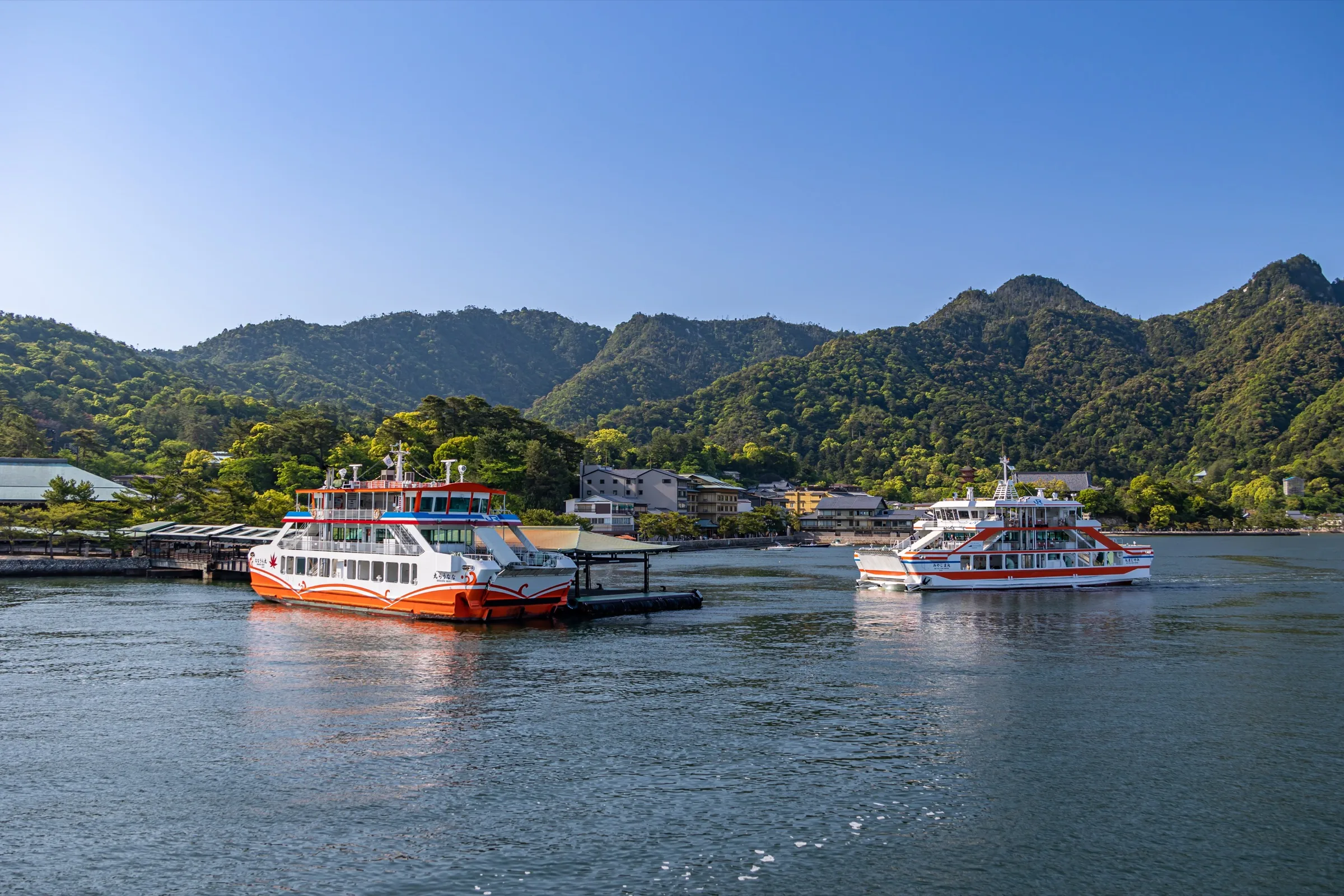 Two JR ferries at Miyajima Pier, with the island’s wooded slope behind the terminal