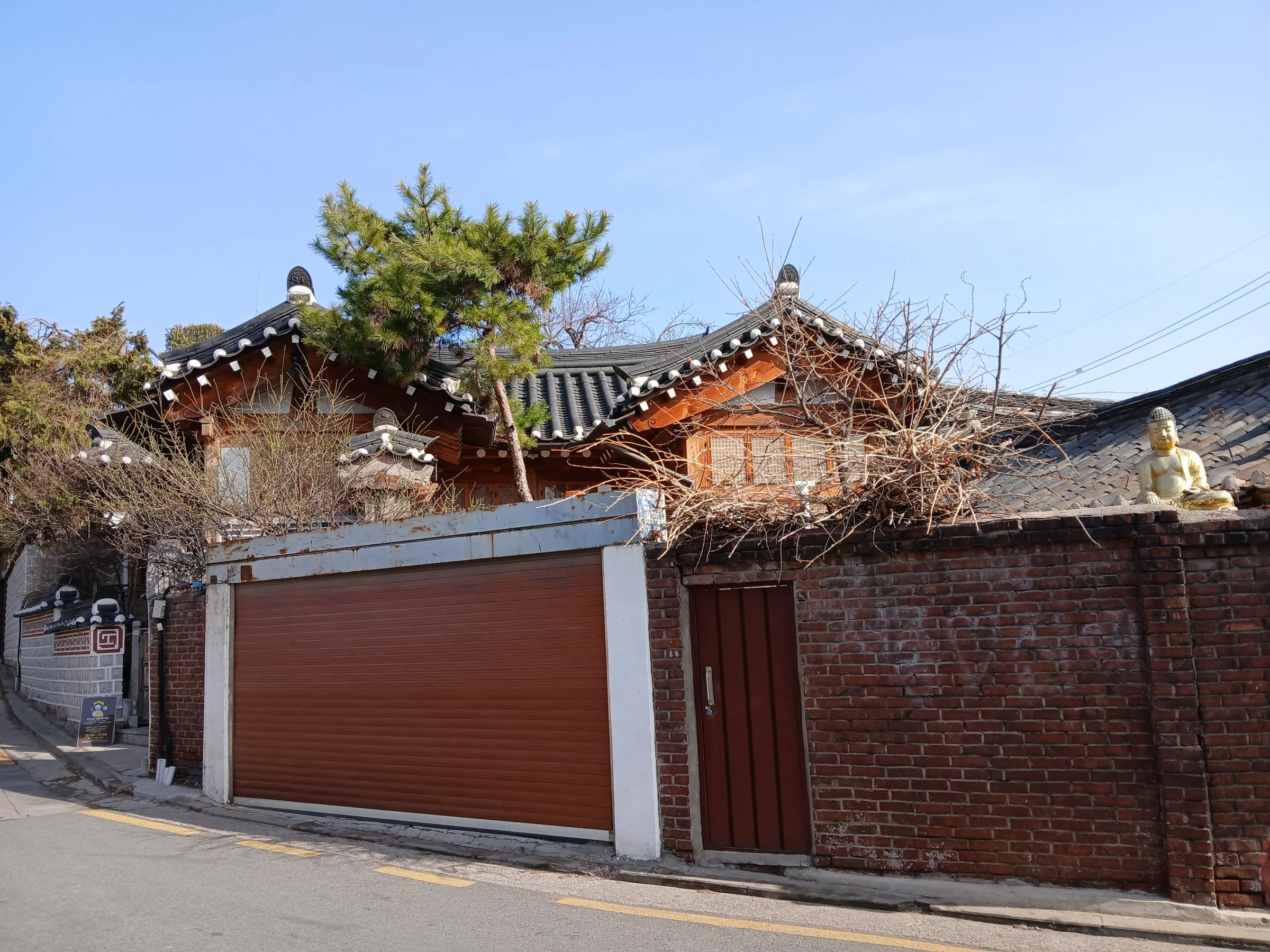 A hanok roof and small Buddha figure seen beyond a brick boundary wall in Bukchon